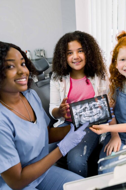 Shot of professional female african dentist and her two little patients, multiethnic shcoolgirls, sitting in dentistry chair, showing digital tablet with jaws and teeth x-ray to camera and smiling Dentist with kids teaching preventative education and dental hygiene